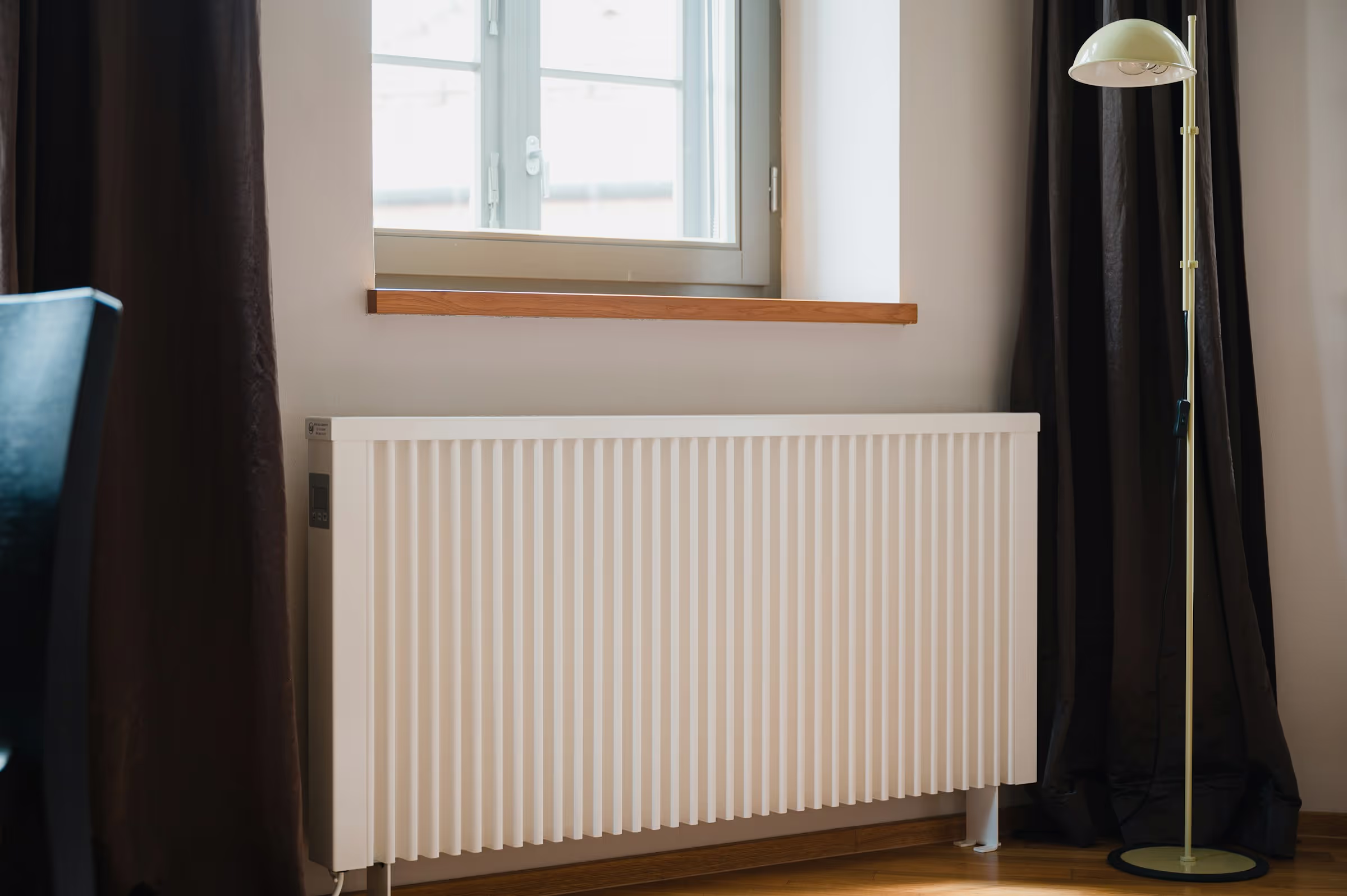Nahaufnahme eines Heizkörpers unter einem Fenster mit Stehlampe und dunklen Vorhängen am Tag.